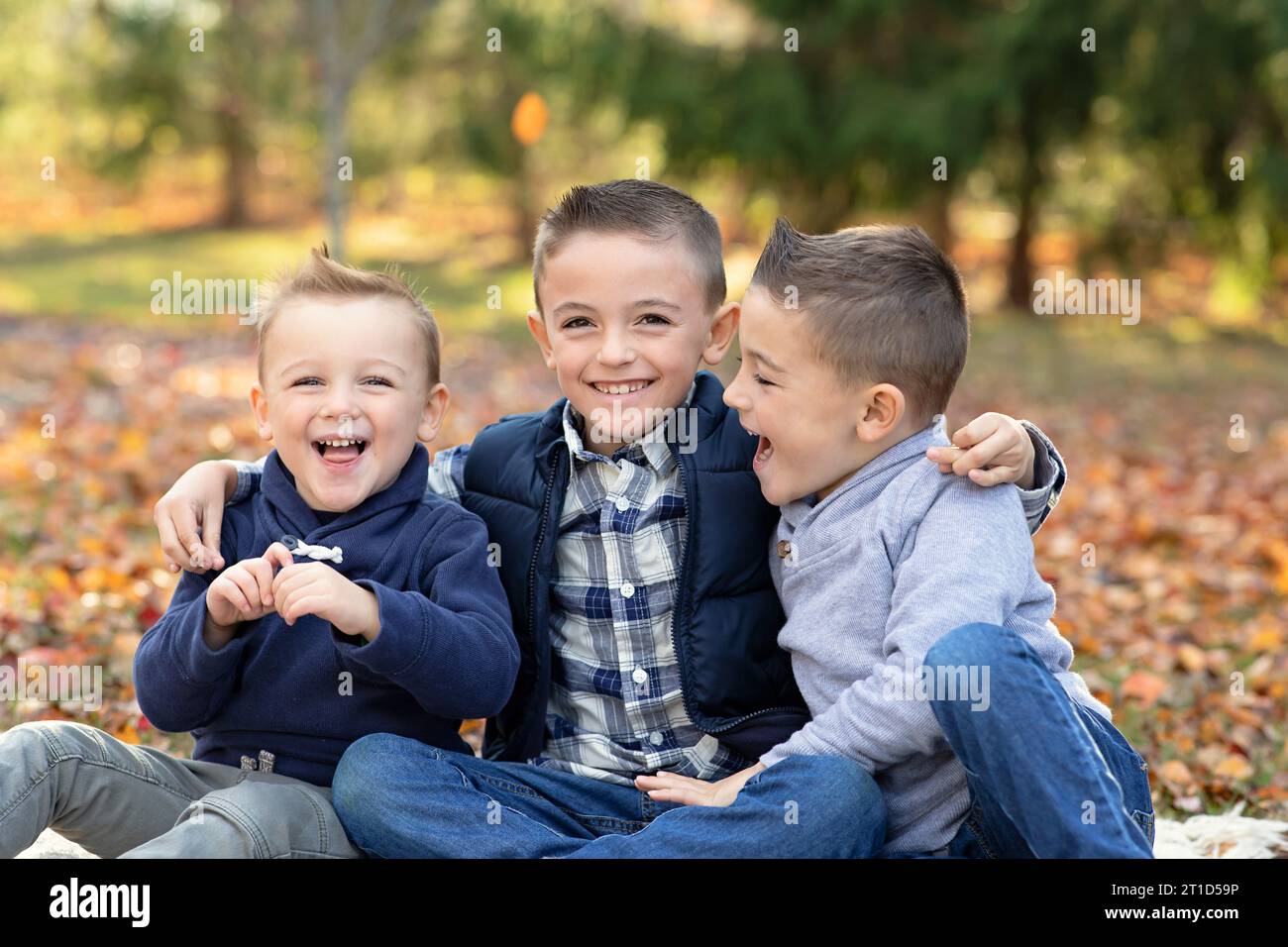 Tre fratelli, 4-10, ridendo, abbracciati, seduti tra le foglie autunnali Foto Stock