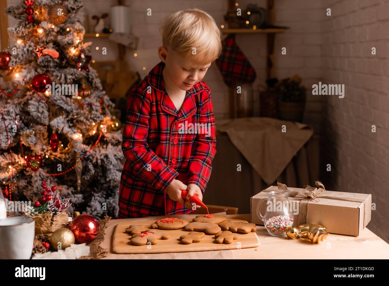 Il ragazzo biondo in pigiama rosso decora i biscotti natalizi con glassa in cucina. Foto Stock