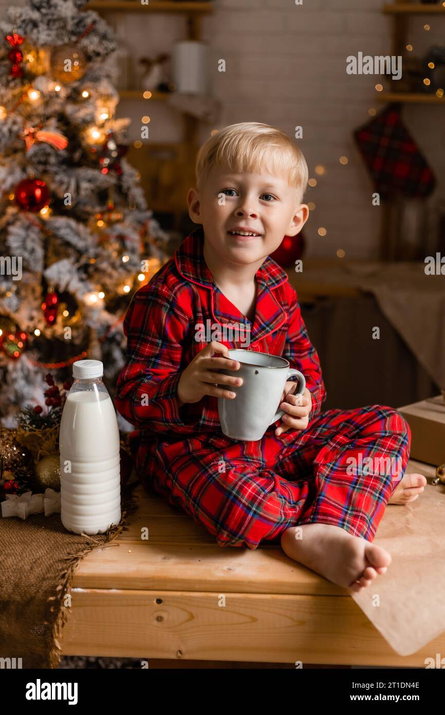 Il ragazzo biondo in pigiama rosso beve latte nella cucina di Natale. Colazione salutare Foto Stock