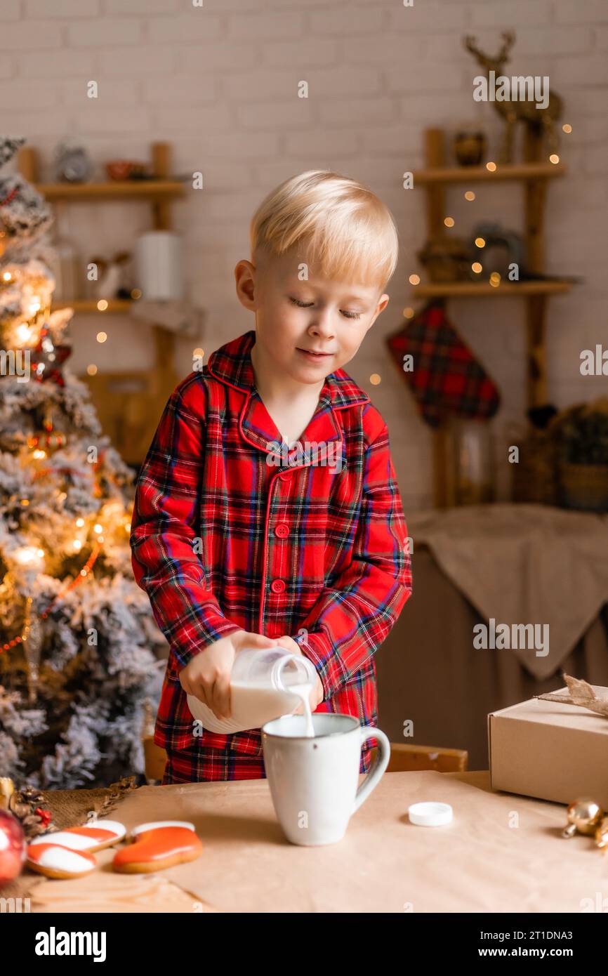 Il ragazzo biondo in pigiama rosso beve latte nella cucina di Natale. Colazione salutare Foto Stock