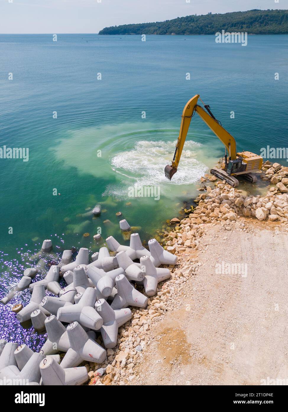 Un escavatore costruisce diligentemente un molo o un frangiflutti nel mare, il suo potente braccio che si estende dalla riva, creando una struttura resiliente in mezzo Foto Stock
