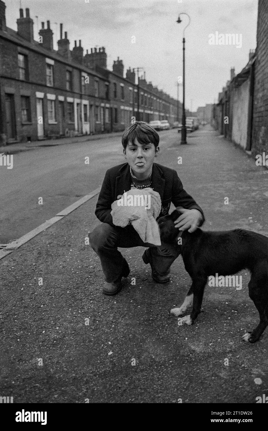 Ragazzo che indossa l'uniforme scolastica e si mette la lingua mentre accarezza il suo cane, foto scattata durante la slum clearance, St Ann's, Nottingham. 1969-1972 Foto Stock