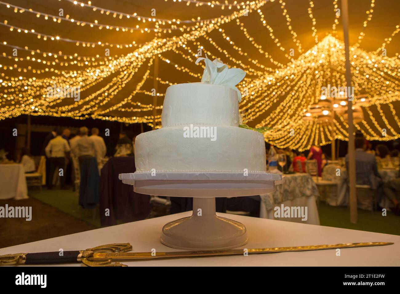 Torta nuziale fondente sul tavolo con la spada nella sala banchetti. Tenda decorata con ghirlande di LED sullo sfondo Foto Stock