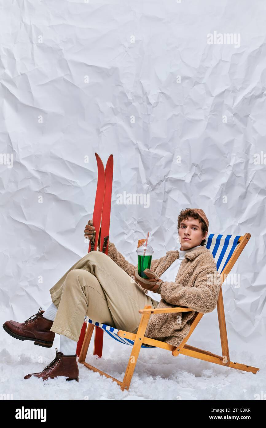 uomo alla moda con un caldo vestito seduto su una sedia a sdraio con un cocktail caldo e un drink in uno studio innevato Foto Stock
