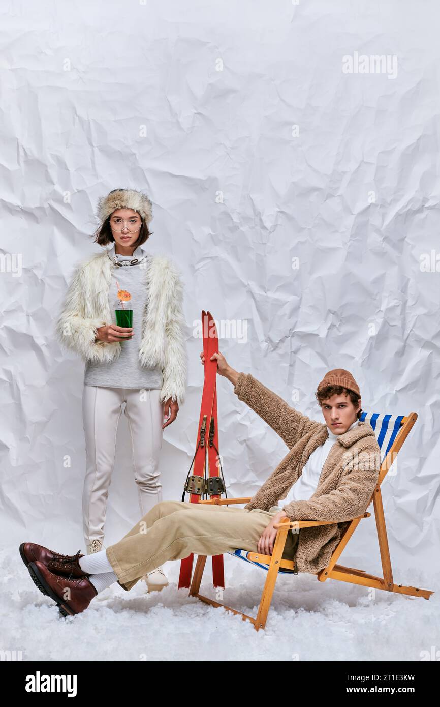 uomo elegante con sci seduto su una sedia a sdraio vicino a una donna asiatica con un cocktail caldo sulla neve in studio Foto Stock