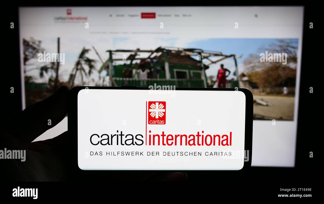 Persona che possiede uno smartphone con il logo dell'agenzia di aiuti Caritas International davanti al sito Web. Concentrarsi sul display del telefono. Foto Stock