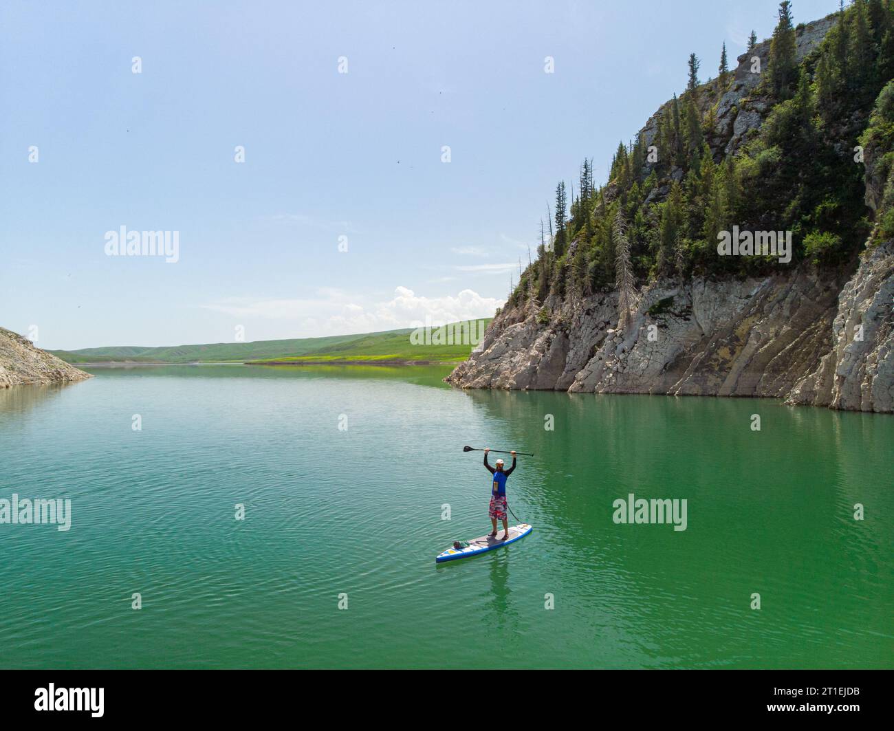Un uomo barbuto nuota su una tavola SUP su un lago tra le rocce. In piedi pagaiando. Lago di montagna con acqua azzurra Foto Stock