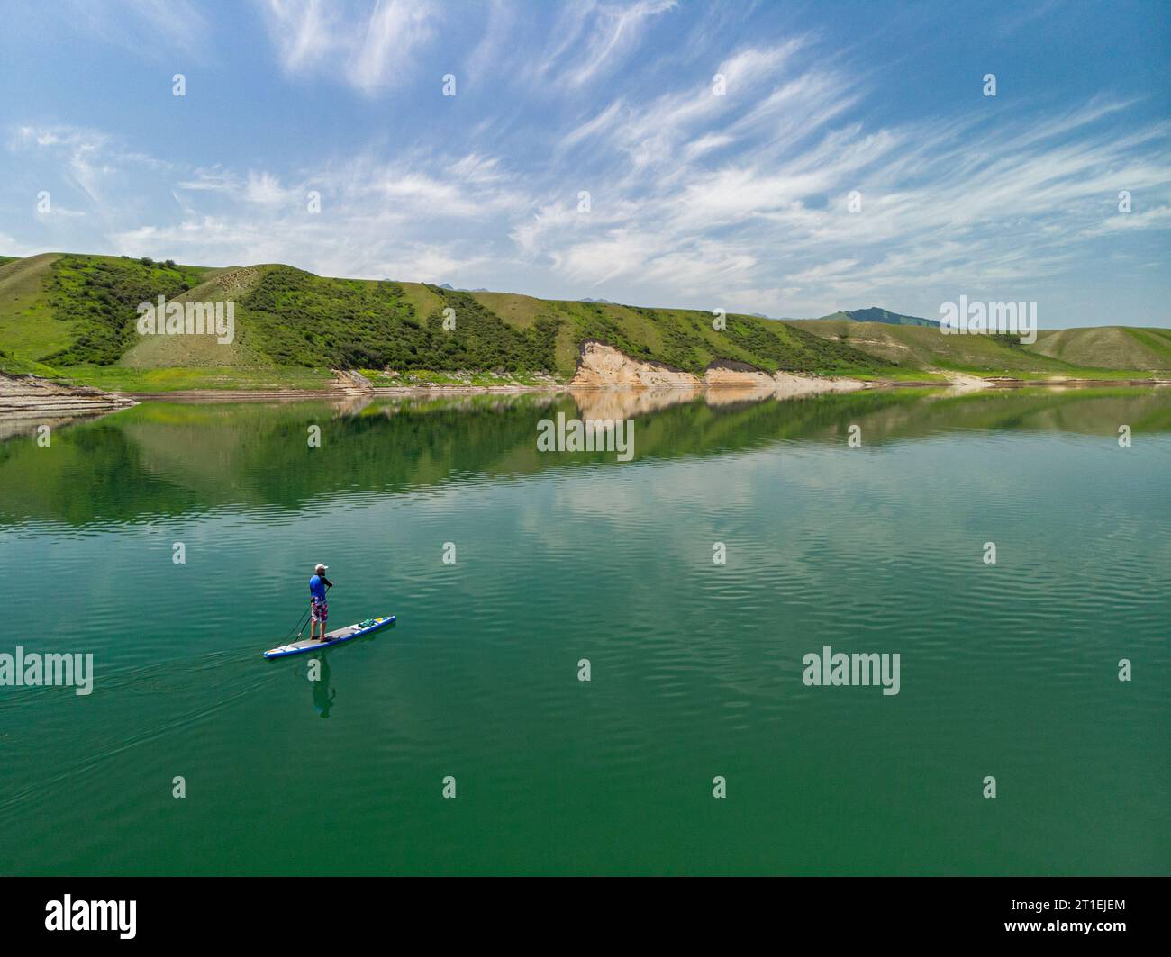 Un uomo barbuto nuota su una tavola SUP su un lago tra le rocce. In piedi pagaiando. Lago di montagna con acqua azzurra Foto Stock