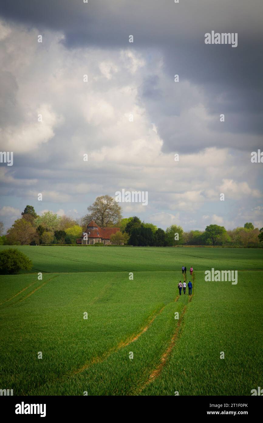 Una scena paesaggistica con un piccolo gruppo di persone che camminano lungo un sentiero in un grande campo, verso una piccola chiesa del villaggio in lontananza. Foto Stock