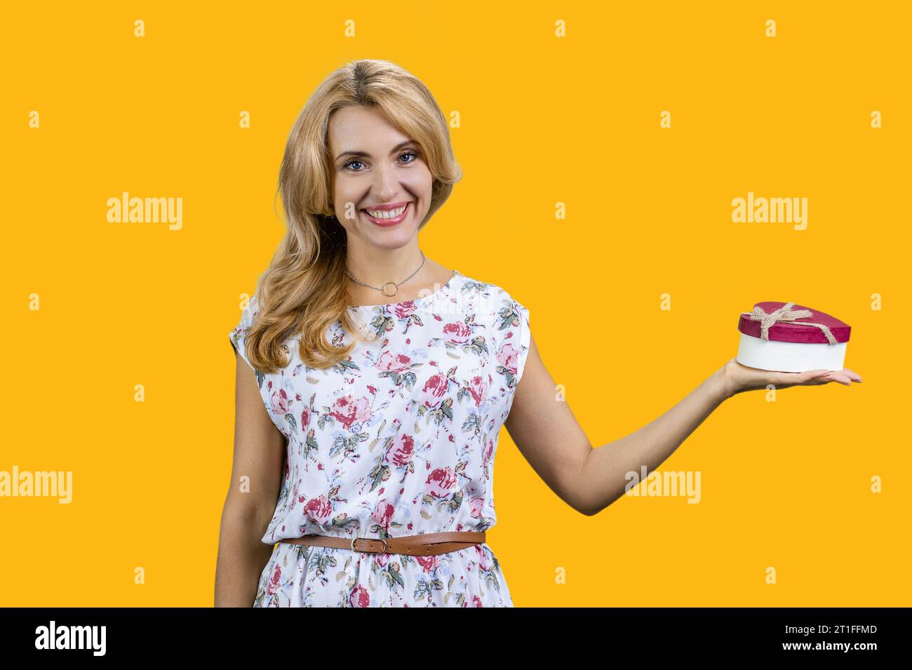 Donna matura bionda con una confezione regalo a forma di cuore sulla mano sinistra. Isolato su giallo. Foto Stock