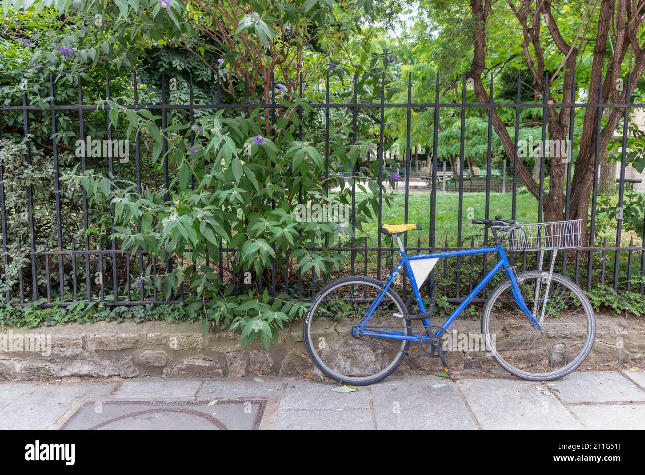 Una bicicletta blu è incatenata alle ringhiere di un parco a Parigi, in Francia. Gli alberi e i cespugli nel parco passano attraverso le ringhiere sul marciapiede. Foto Stock