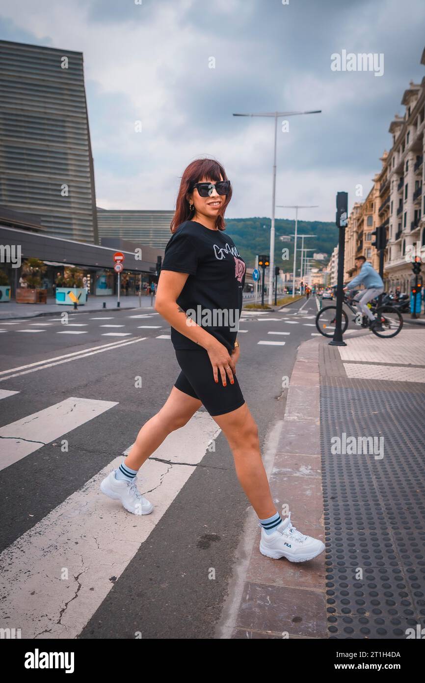 Stile di vita in città. Una giovane bruna con occhiali da sole, shorts neri, una T-shirt nera e sneakers bianche. Attraversa una zebra nella città successiva Foto Stock