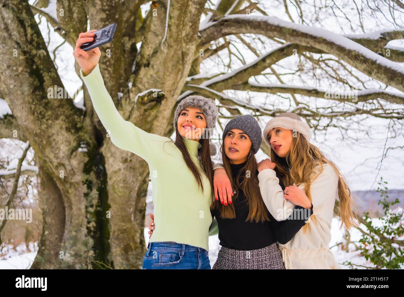 Stile di vita invernale, tre bellissimi amici caucasici che scattano un selfie con il telefono nella neve sotto un albero, vacanze nella natura Foto Stock