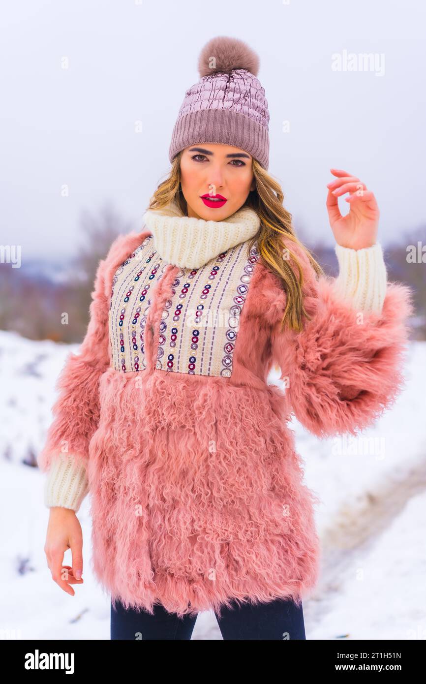 Ritratto di una giovane modella bionda che indossa una giacca di pelliccia rosa, stivali invernali e un cappello viola sulla neve. Stile di vita Foto Stock