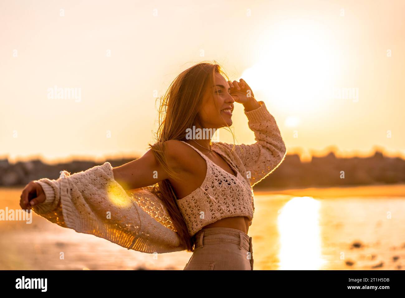 Stile di vita estivo. Una giovane donna bionda caucasica con un maglione bianco corto in lana al tramonto sulla spiaggia. A braccia aperte in riva al mare Foto Stock