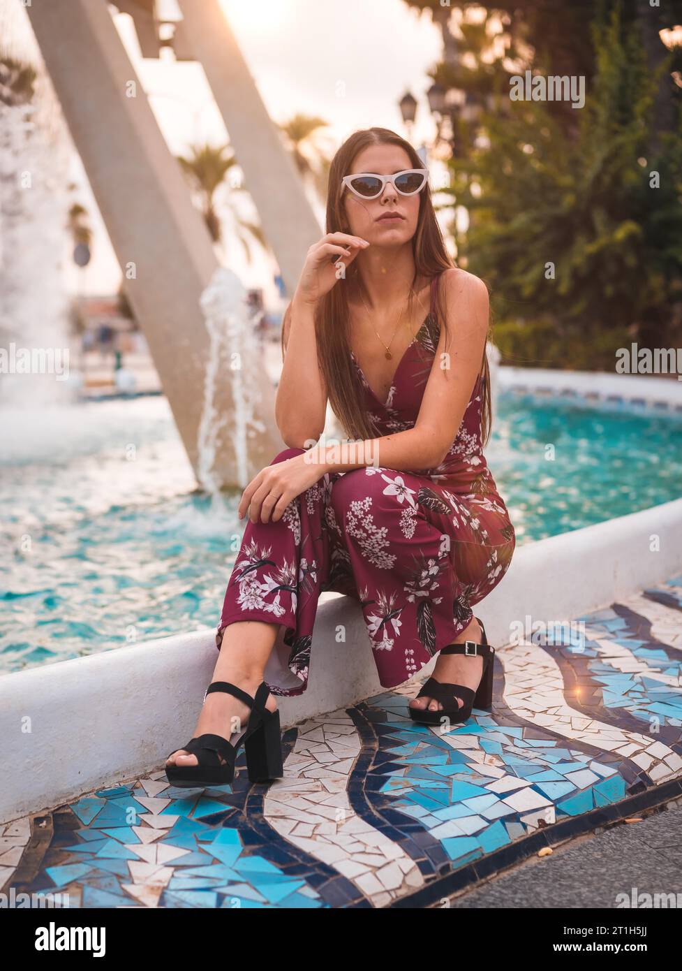 Ritratto di una giovane bruna in abito floreale marrone e moderni occhiali da sole bianchi che si godono l'estate nell'ora d'oro in una fontana della città Foto Stock