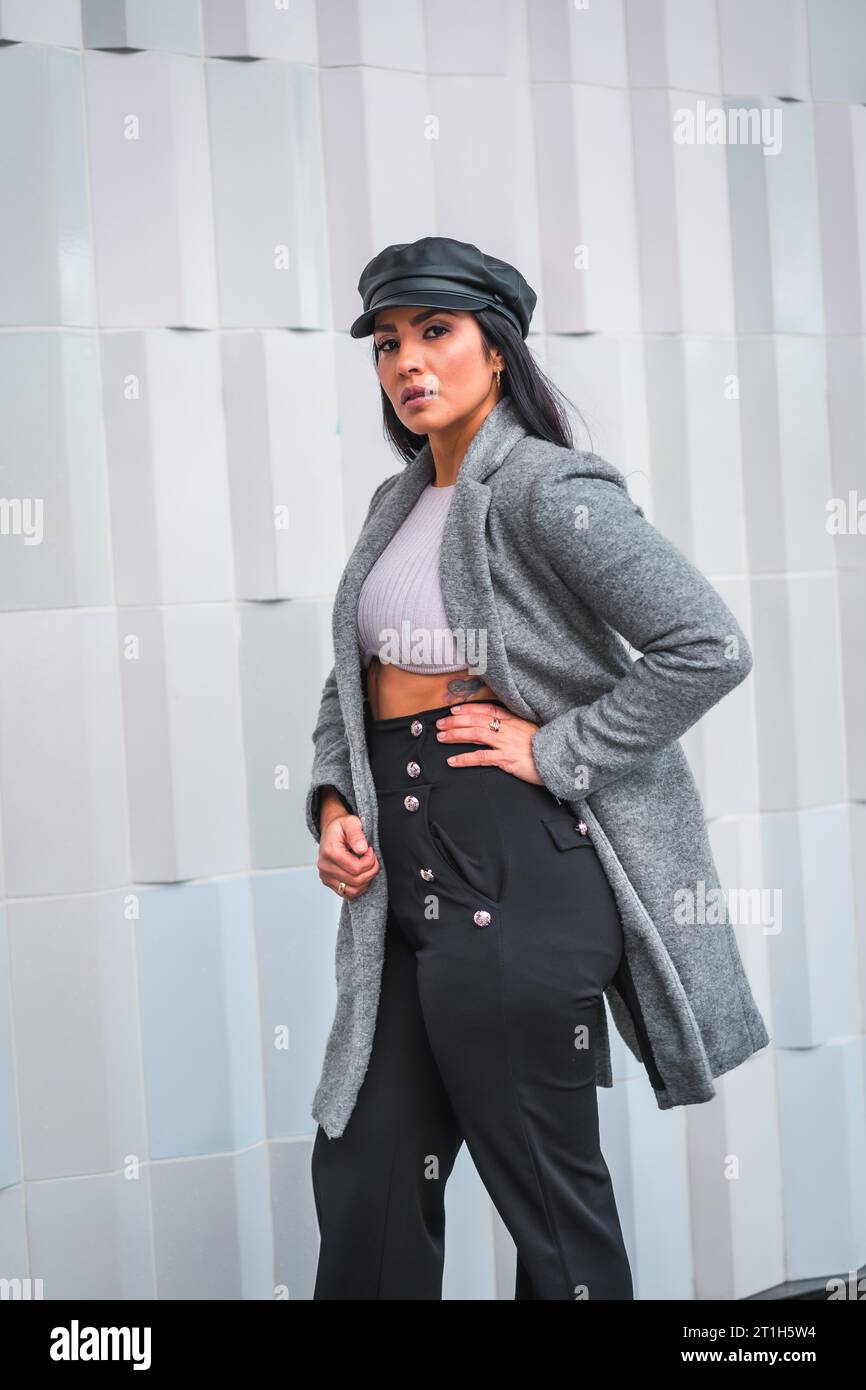 Una giovane bruna latina con un berretto in pelle e un blazer grigio in un ritratto con uno sfondo grigio della città Foto Stock