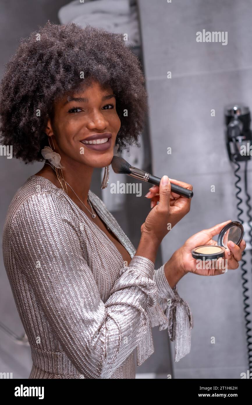 Una giovane ragazza nera con i capelli afro che si è messa il trucco prima della festa con un sorriso. Festa esclusiva, festa vip, abito da sera grigio Foto Stock