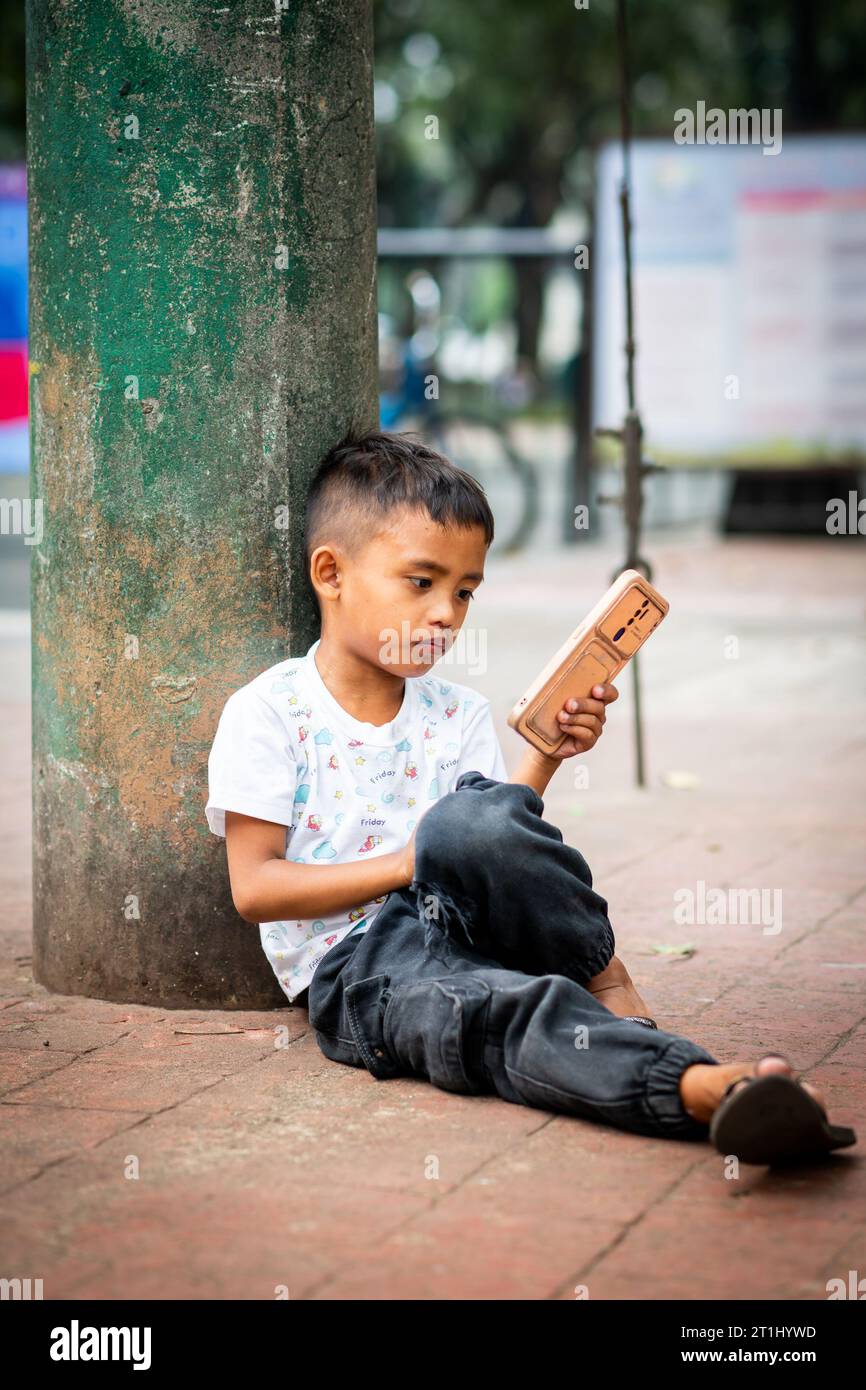 Un giovane filippino gode di qualcosa sul suo cellulare seduto fuori da Rizal Park, Manila, Filippine. Foto Stock