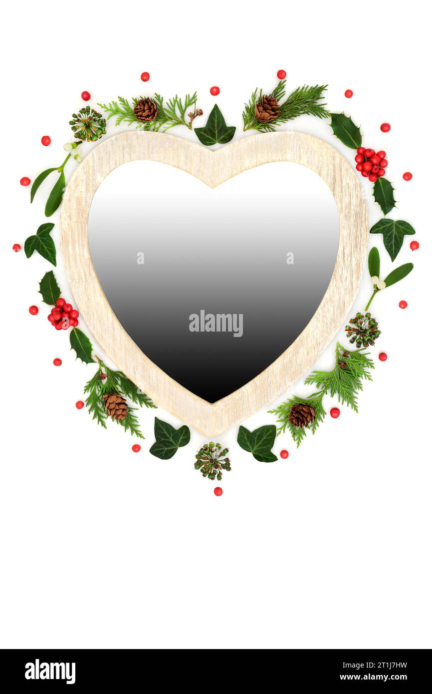 Corona natalizia a forma di cuore con holly, vischio e vegetazione invernale festosa. Simbolo tradizionale per le feste su sfondo bianco. Foto Stock