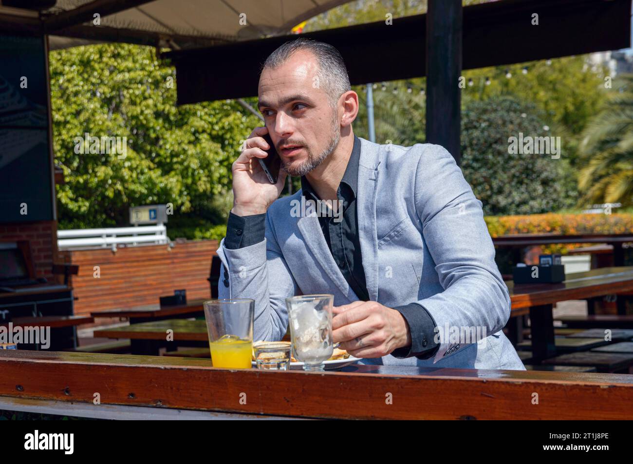 Uomo d'affari caucasico adulto, con abiti eleganti, seduto con il telefono in mano, finendo il cappuccino, ristorante all'aperto, colazione in mo Foto Stock