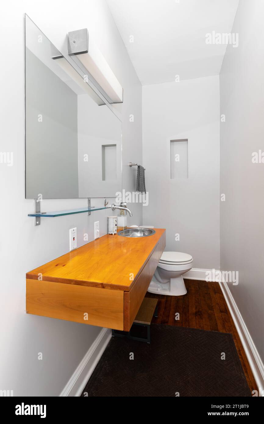 Un piccolo bagno con armadio galleggiante in legno, pavimento in legno duro, lavandino e rubinetto in acciaio inossidabile. Foto Stock