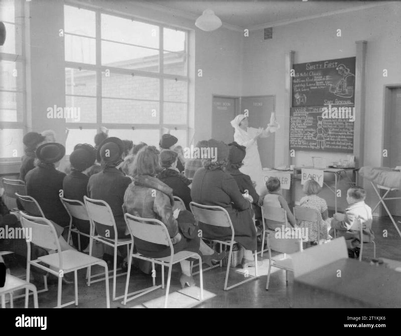 Asilo nido a Tottenham, Londra, Inghilterra, 1940 Un infermiere visualizza una bottiglia di latte a una classe come parte di un tempo di guerra clinica per le madri detenute presso il parco comunale di corsia di asili nido in Tottenham. Sulla lavagna dietro di lei, informazioni sulla cucina di guerra viene visualizzato, compresi suggerisce una colazione a base di uova strapazzate e una cena a base di carni di montone e di rognone. La Blackboard mette anche in evidenza il fatto che non ci sono ante-natal cliniche a West Green Road, Signoria Lane e Park Lane. Foto Stock