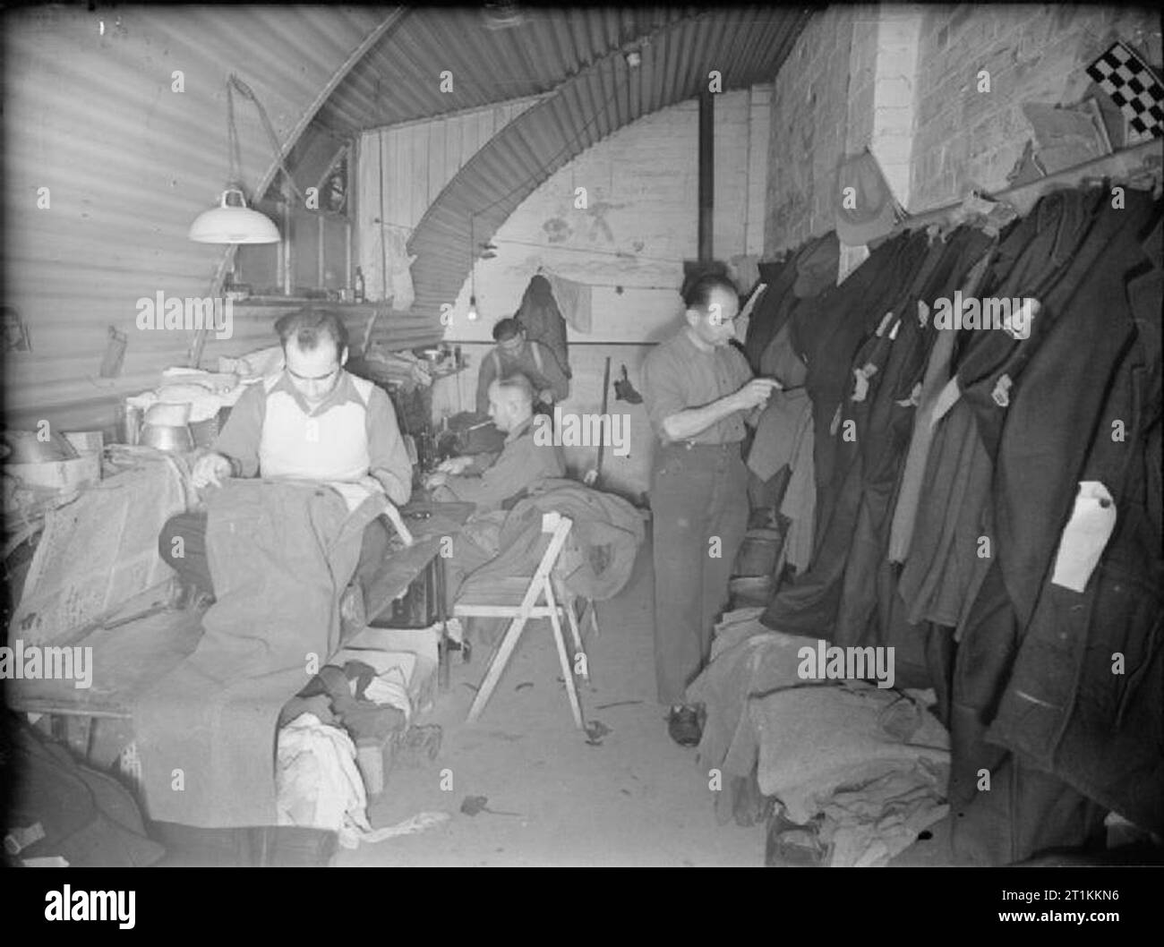 Prigionieri di guerra tedeschi in Gran Bretagna - La vita quotidiana in un tedesco POW Camp, UK, 1945 prigionieri di guerra tedeschi mend vestiti nella sartoria di un accampamento di PRIGIONIERO DI GUERRA, da qualche parte in Gran Bretagna. Tute sono appese in alto a destra della fotografia. Un uomo è utilizzando una macchina da cucire. La parete posteriore del Nisseno hut utilizzato come la sartoria ha un murale di un sommergibile disegnata su di esso. Foto Stock