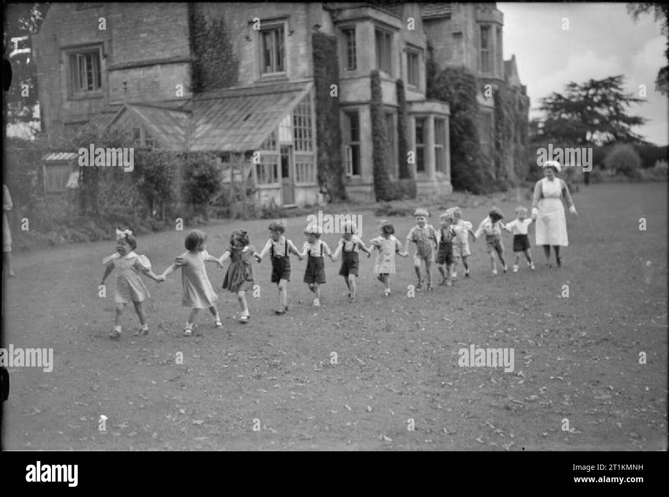 La vita al Vivaio Querns (l'Arthur James Balfour Home), Gloucestershire, ottobre 1942 bambini tenere mani e formano una linea come essi camminare attraverso l'erba in motivi di Querns vivaio nel Gloucestershire. I loro sentieri infermiere lungo dietro, cercando di tenere il passo con il loro gioco di Caterpillar. Foto Stock