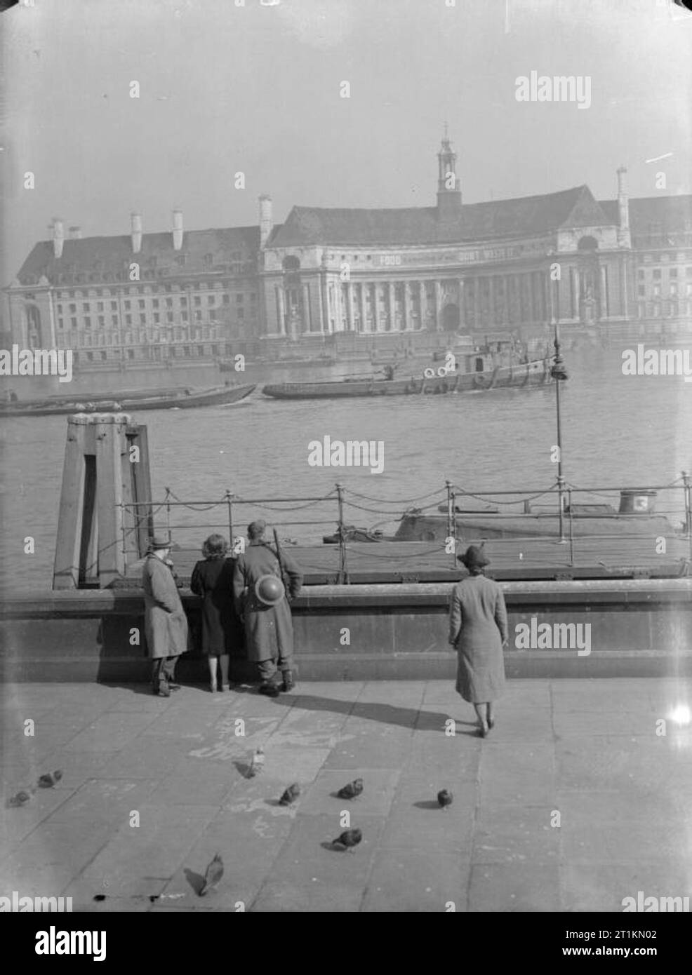 Londra in primavera del 1941- La vita quotidiana a Londra in Inghilterra un soldato e una tre civili pausa nel loro cammino lungo il Victoria Embankment per guardare una chiatta viaggio verso il fiume Tamigi passato County Hall. A pochi piccioni può essere visto in primo piano, cercando le briciole. Foto Stock