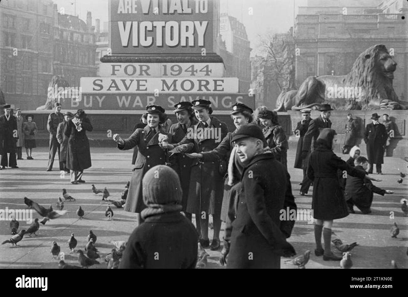 Londra nel quinto anno di guerra - La vita quotidiana in London, 1944 membri della donne ausiliario della Air Force e i bambini sono tra quelli di alimentazione del piccioni in Trafalgar Square a Londra. In background, alla base della colonna di Nelson, è coperta in guerra poster di risparmio ed uno di Trafalgar Square i Lions possono anche essere visto. Foto Stock