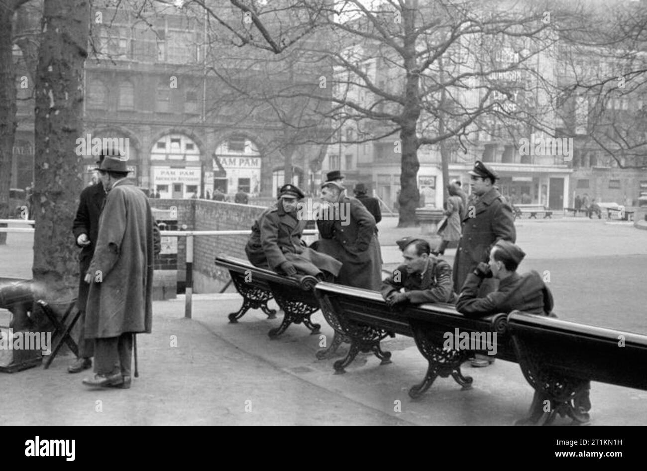 Londra nel quinto anno di guerra - La vita quotidiana in London, England, Regno Unito, 1944 Un gruppo di inglesi e americani il personale di servizio chat assieme sulle panchine nel cuore di Leicester Square. Dietro di loro un grande 'American Restaurant' chiamato 'Pam Pam'. A destra della fotografia, dietro l'albero, il Leicester Square Theatre può essere visto. Tra gli uomini e il ristorante, il mattone entrata di un raid aereo riparo può essere visto. Altri pedoni fanno la loro strada attraverso la piazza come andare circa le loro attività quotidiane. Foto Stock