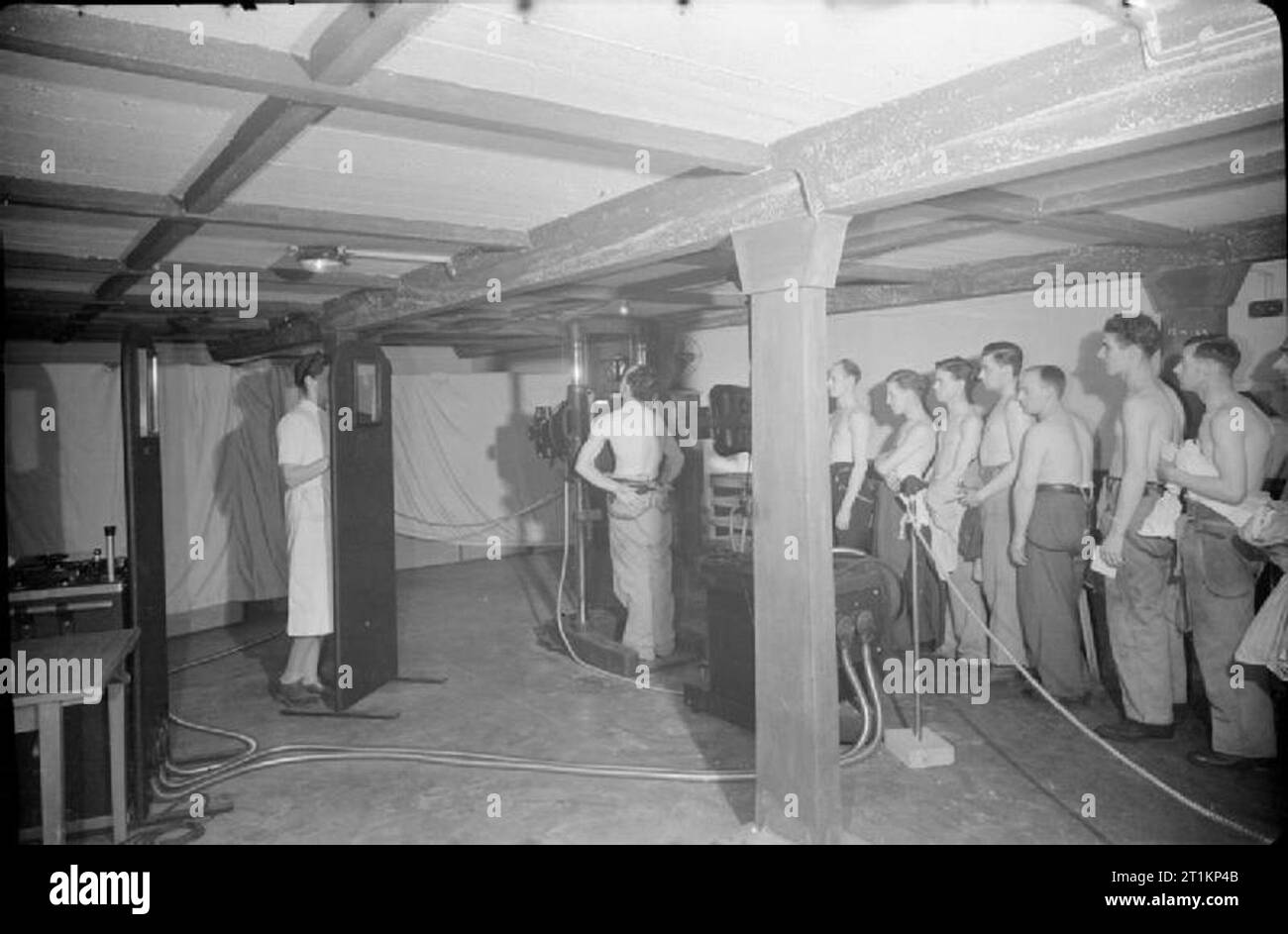 Nuova guerra si apre sulla parte anteriore di fabbrica- X-Ray Screening per la tubercolosi, Londra, Inghilterra, 1943 Una vista a raggi x in corso in una clinica di fortuna nel rifugio in mensa di questa fabbrica, da qualche parte nella periferia di Londra. Un lavoratore è stato detto di prendere un respiro profondo un tenerlo premuto mentre i raggi x è preso dalla tabella di controllo operatore (non visibili nella foto). L'infermiera che posizioni di persone nel posto giusto per loro x-ray, sorge dietro uno schermo di protezione, al fine di evitare una eccessiva esposizione. Altri lavoratori in coda, pronto per il loro turno per essere radiografato. Foto Stock