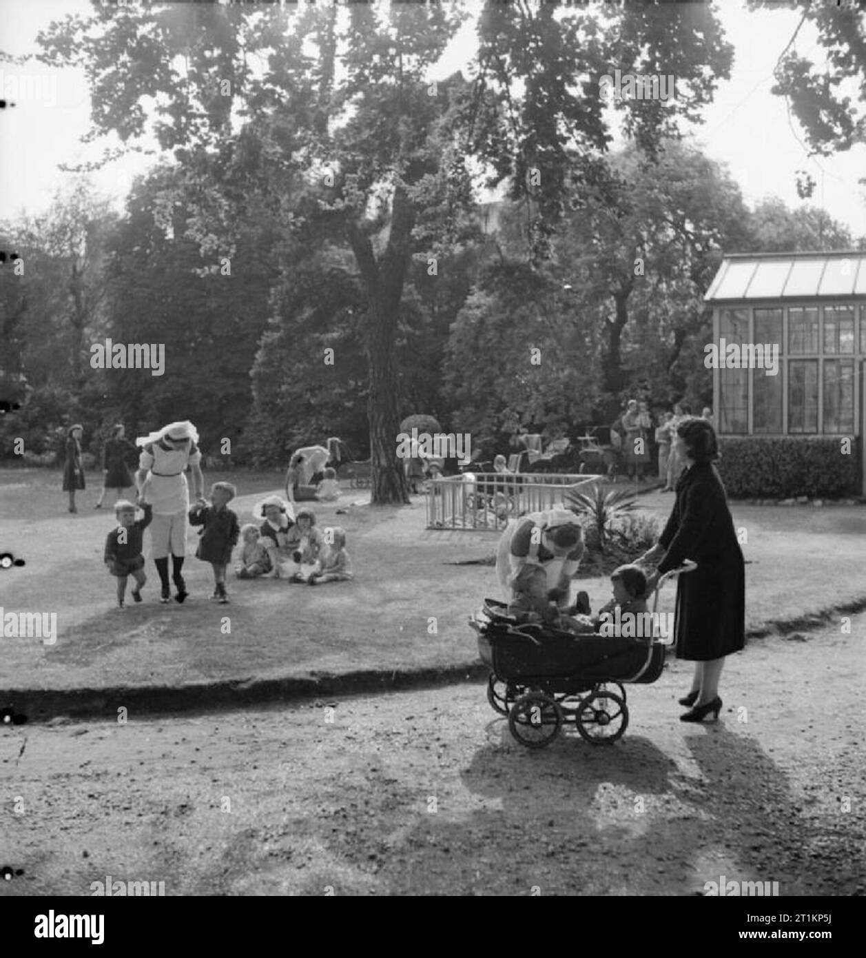 Nursery per le mamme che lavorano- il lavoro della Pietra Focaia Strada Verde vivaio, Birmingham, 1942 La signora Sullivan raccoglie i suoi figli dalla pietra focaia verde vivaio di strada alla fine della sua giornata di lavoro. Un infermiere si infila 18 mese vecchio Giovanni nella PRAM accanto alla sua 3 e una metà di anni di Suor Maureen. Dietro di loro, un altro infermiere conduce due figli a trovare la loro madre e gli altri bambini giocare sul prato di sera il sole. Foto Stock