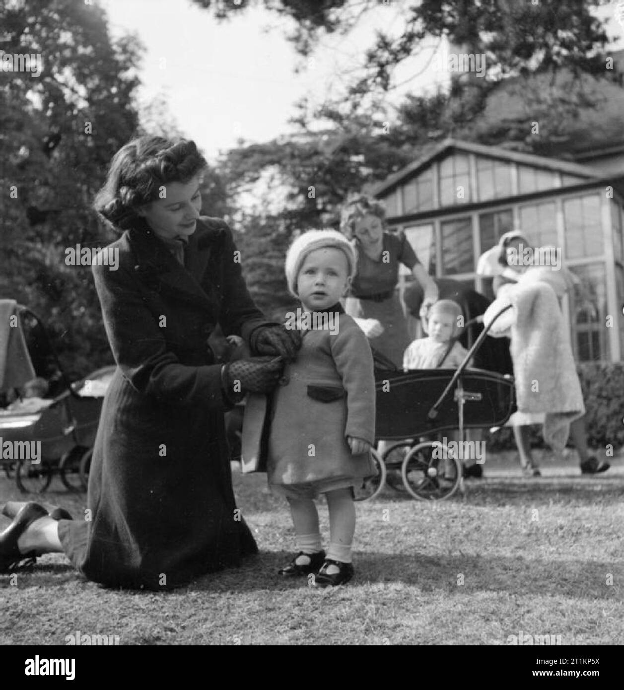 Nursery per le mamme che lavorano- il lavoro della Pietra Focaia Strada Verde vivaio, Birmingham, 1942 20 enne ra Ann Donovan serra i pulsanti sul suo figlio Michael del mantello e come essa raccoglie lui dal giardino del vivaio. Michael è di 16 mesi. Onorevole Donovan ha appena completato una giornata di lavoro in una fabbrica di munizioni. Dietro di loro un altro madre può essere visto la raccolta di sua figlia in una carrozzina e un infermiere porta un altro bambino a una diversa parte del giardino. Foto Stock