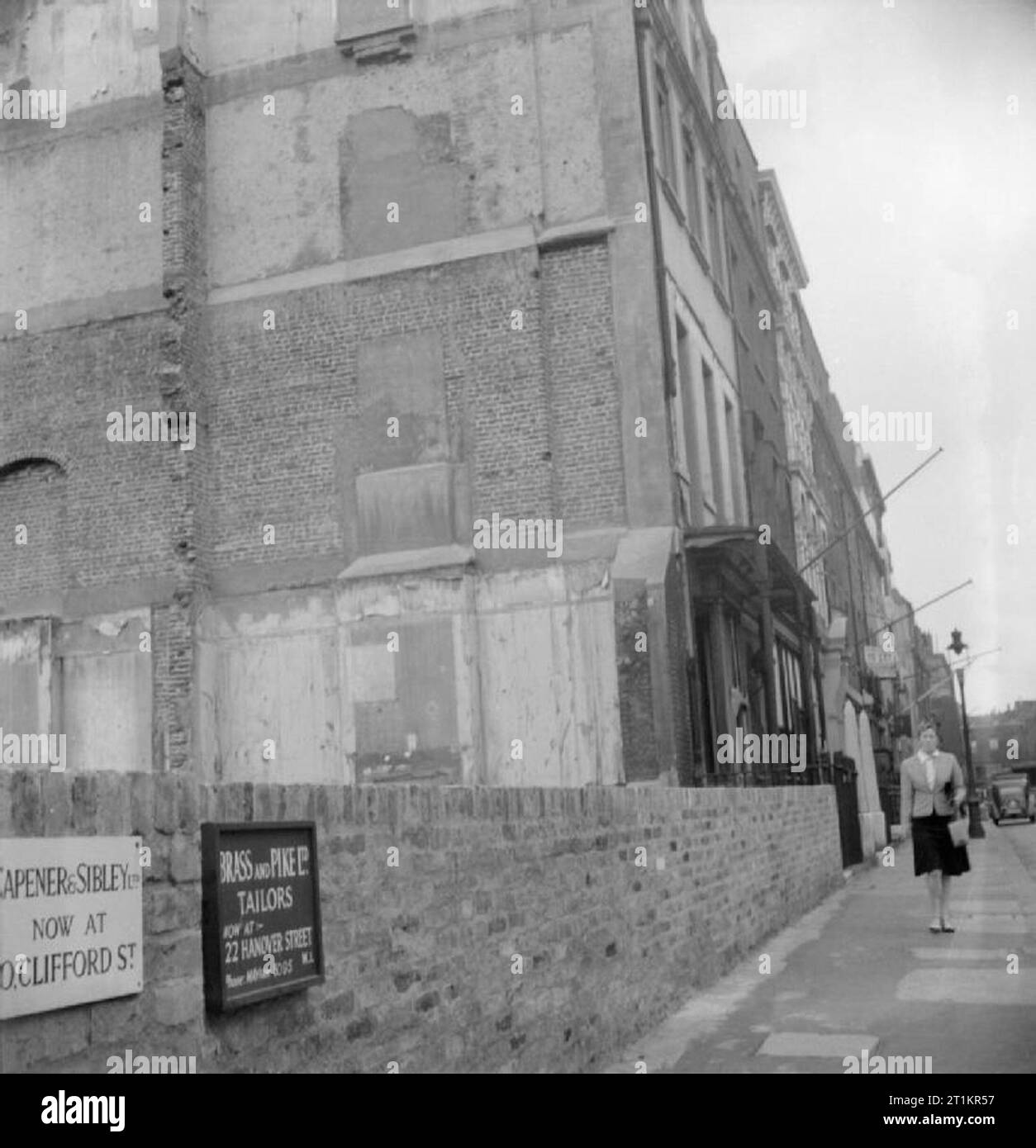 Savile Row- Sartoria presso Henry Poole e Co. di Londra, Inghilterra, Regno Unito, 1944 Una donna cammina giù Savile Row, passato lo spazio lasciato case dove una volta sorgeva, prima che fossero distrutte in un raid aereo. Il sito è ora sede di un'emergenza serbatoio acqua, nascosto dietro la parete. Chiaramente visibili sono due schede consulenza ai clienti dei nuovi indirizzi di 'Capener e Sibley Ltd.", (ora alla nuova sede di Clifford Street), e 'l'ottone e il luccio Sarti Ltd. (ora a 22 Hanover Street). Foto Stock