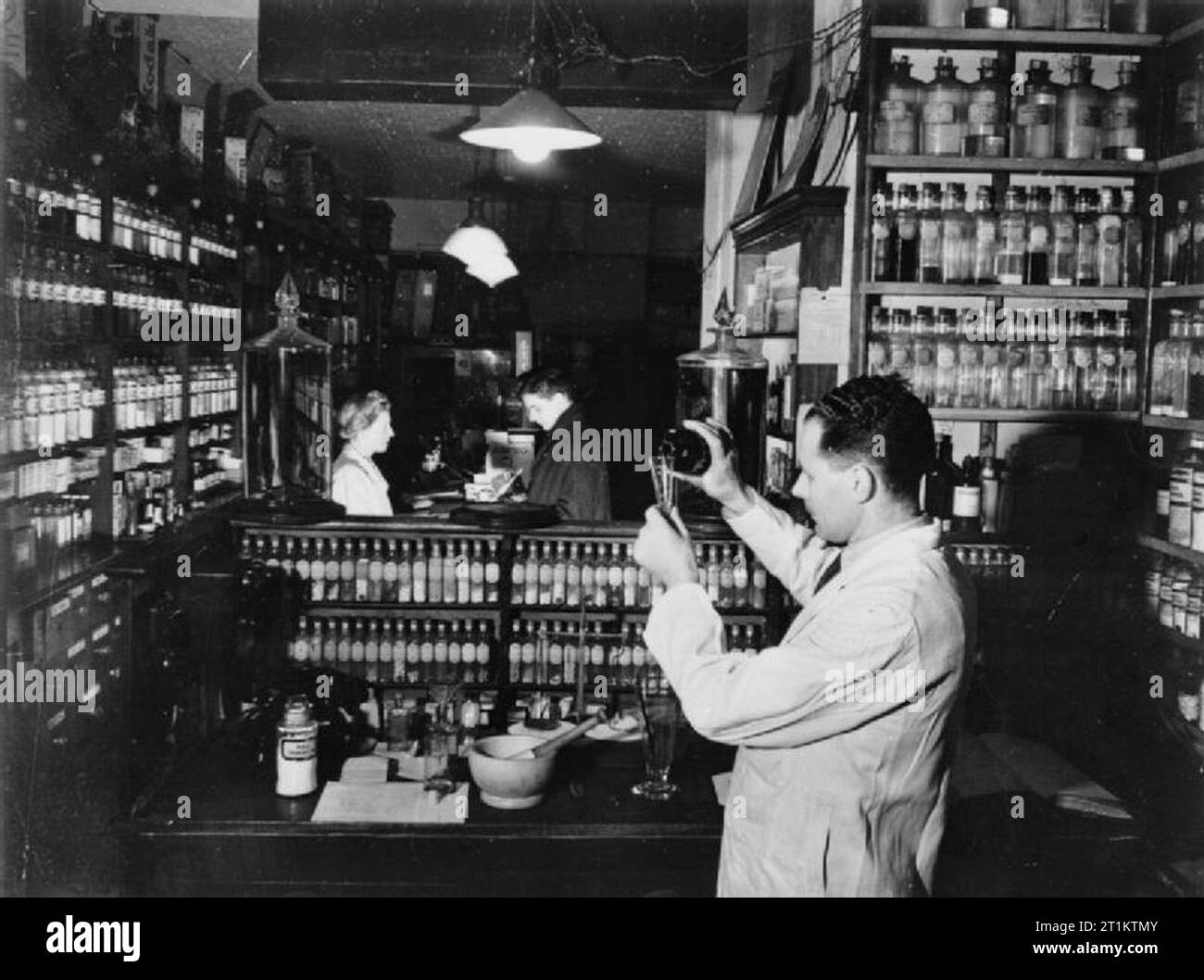 Oppio turco per la Gran Bretagna- l'uso di oppio in medicina britannica, UK, 1943 una scena in un punto di vendita al dettaglio farmacia, mostrando il chimico che compongono una prescrizione del medico da galenicals ricevuto da una farmacia di fabbricazione. Galenicals sono estratti concentrati da cui prescrizioni può essere realizzato e, secondo la didascalia originale, arrivano al farmacista 'come tintura di oppio, concentrato tintura, estratto liquido, morbido estratto, estratto secco o un composto di tintura con canfora e semi di anice'. Un veterano può essere visto presso il contatore, chiedendo una prescrizione dall'assistente. La captio originale Foto Stock
