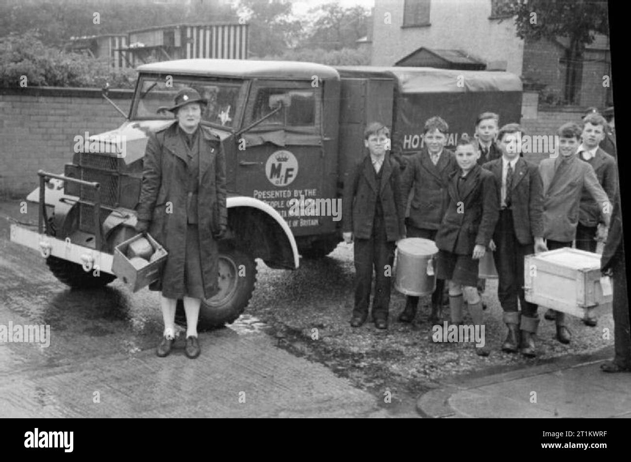 WVS Hot Meals Service- Wartime Food Distribution, Birkenhead, Cheshire, 1942 la signora Jacob si ferma accanto al furgone per una fotografia con un gruppo di bambini che la stanno aiutando a trasportare cibo dal furgone nella loro scuola. La signora Jacob è l'organizzatrice centrale del servizio volontario femminile Birkenhead e porta con sé una scatola di pane. Il Ministero dell'alimentazione è stato presentato dal popolo di Malta. Sembra piovere. Foto Stock