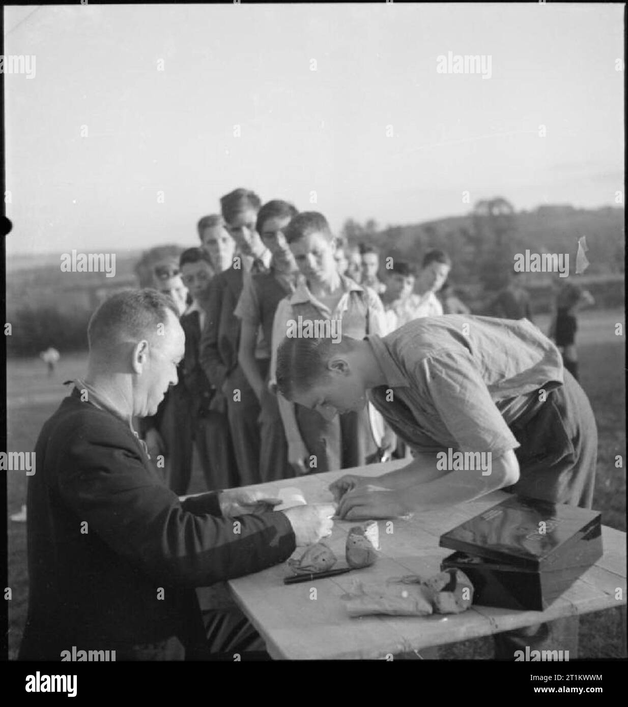 Youth Service Volunteers Help British Farmers- Agricultural Camp at Nunney Catch, Somerset, England, UK, 1943 Un membro del Youth Service Volunteers firma per il suo denaro in tasca durante la parata retributiva al campo agricolo di Nunney Catch. Consegnare i soldi è il direttore del campo, signor Booth, ex ufficiale dell'esercito e della RAF, invalidato dai servizi. I ragazzi aspettano pazientemente di ritirare il loro pagamento settimanale di sei scellini . Foto Stock