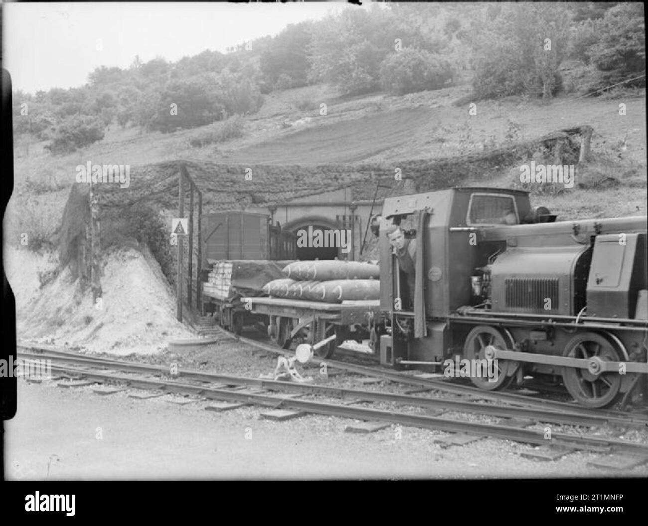 La Royal Navy durante la seconda guerra mondiale Una locomotiva diesel, su una ferrovia a scartamento ridotto, esce da una rivista sotterranea presso il Royal Naval Armament Depot Dean Hill. Il veicolo tira autocarri a pianale piatto e chiusi contenenti munizioni. bombe aeree da 500 libbre possono essere viste sul primo camion a pianale piatto. Notate la rete mimetica sulla via d'ingresso. Foto Stock