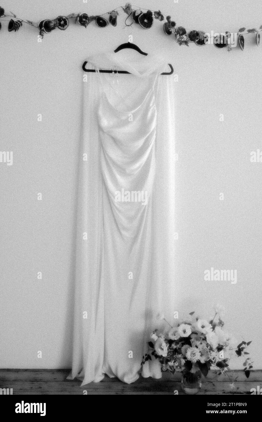 Un elegante abito da sposa bianco esposto su una parete bianca, adornato da un bouquet di fiori Foto Stock