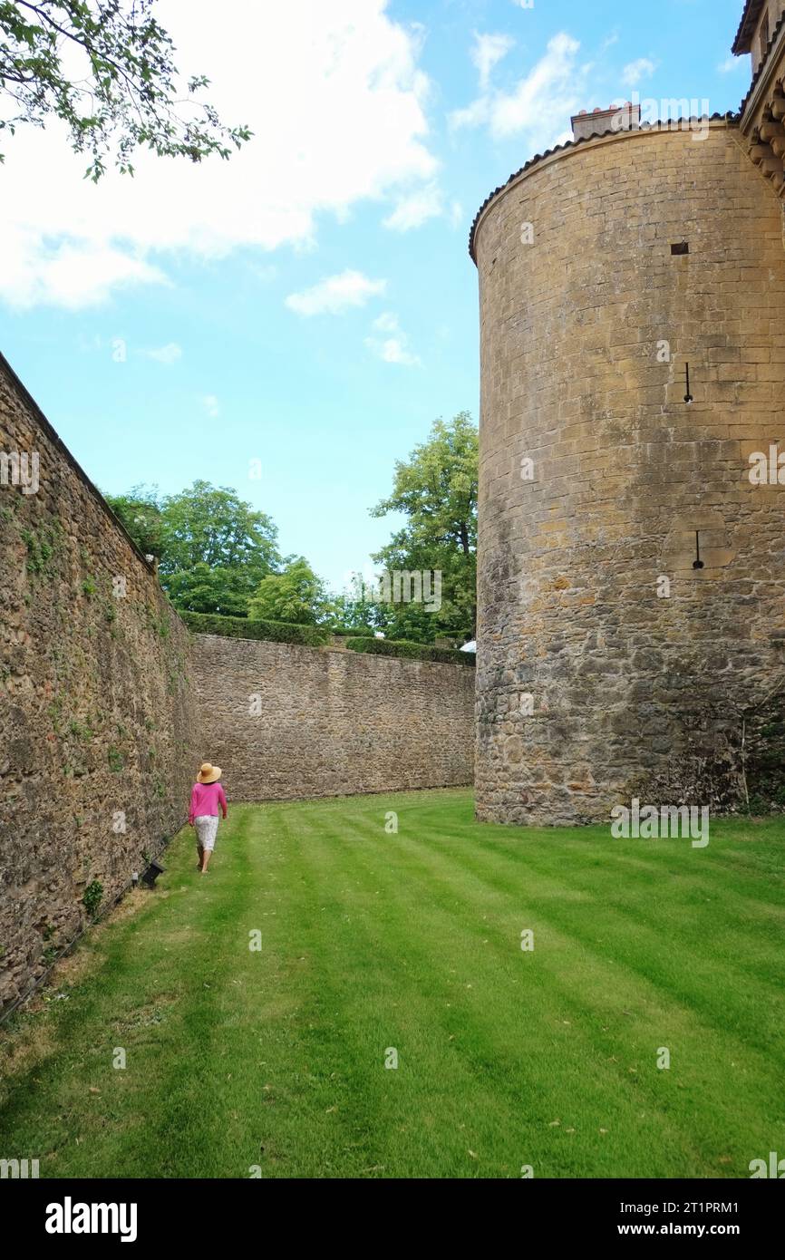 Una donna in un cardigan rosa cammina nel fossato erboso guardando l'architettura e le antiche mura in pietra di Château Bagnols Foto Stock