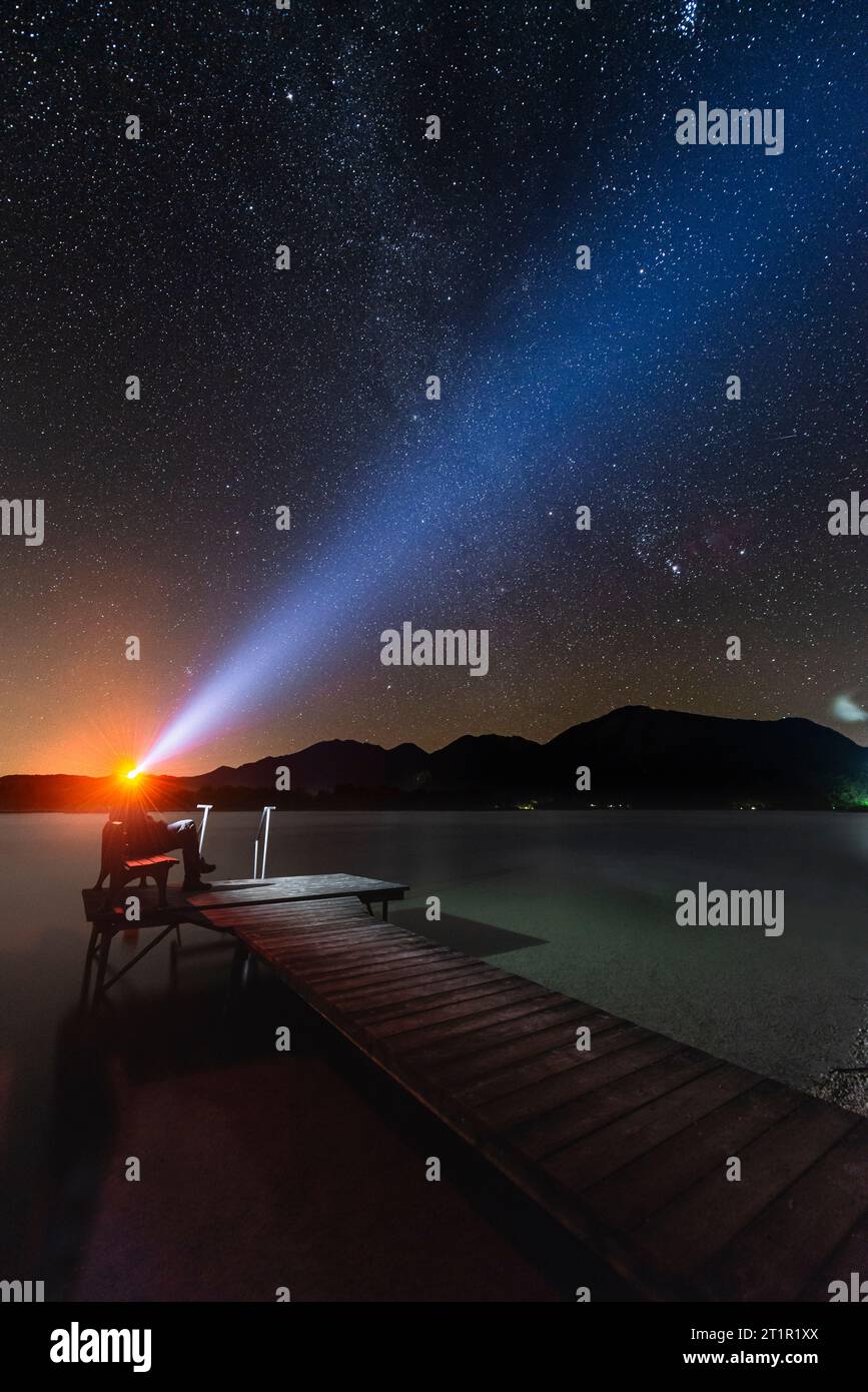 Uomo seduto su una panchina su un molo di legno sulla riva del lago Kochel in Baviera, con un fascio di luce dal suo faro al cielo stellato notturno Foto Stock