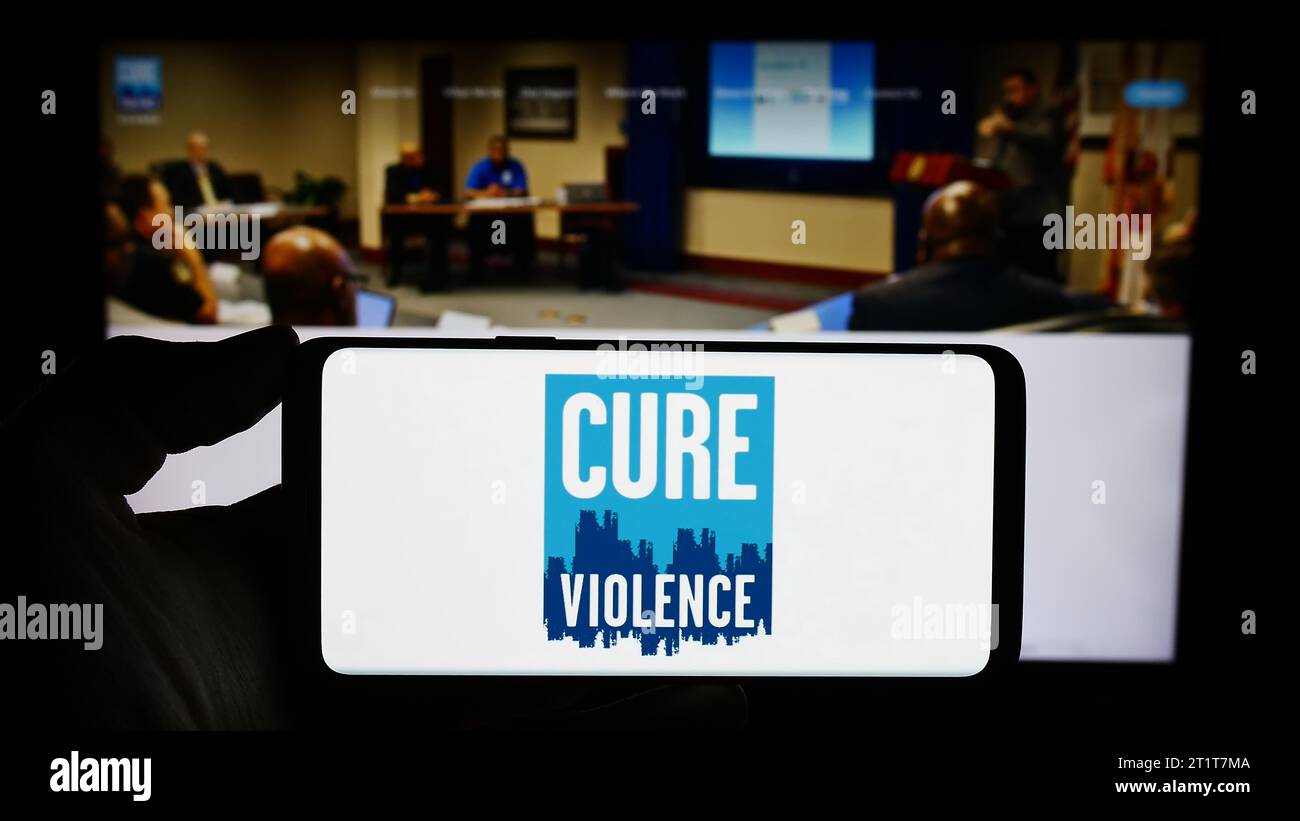 Persona in possesso di un cellulare con il logo dell'organizzazione statunitense cure Violence Global davanti alla pagina web. Concentrarsi sul display del telefono. Foto Stock