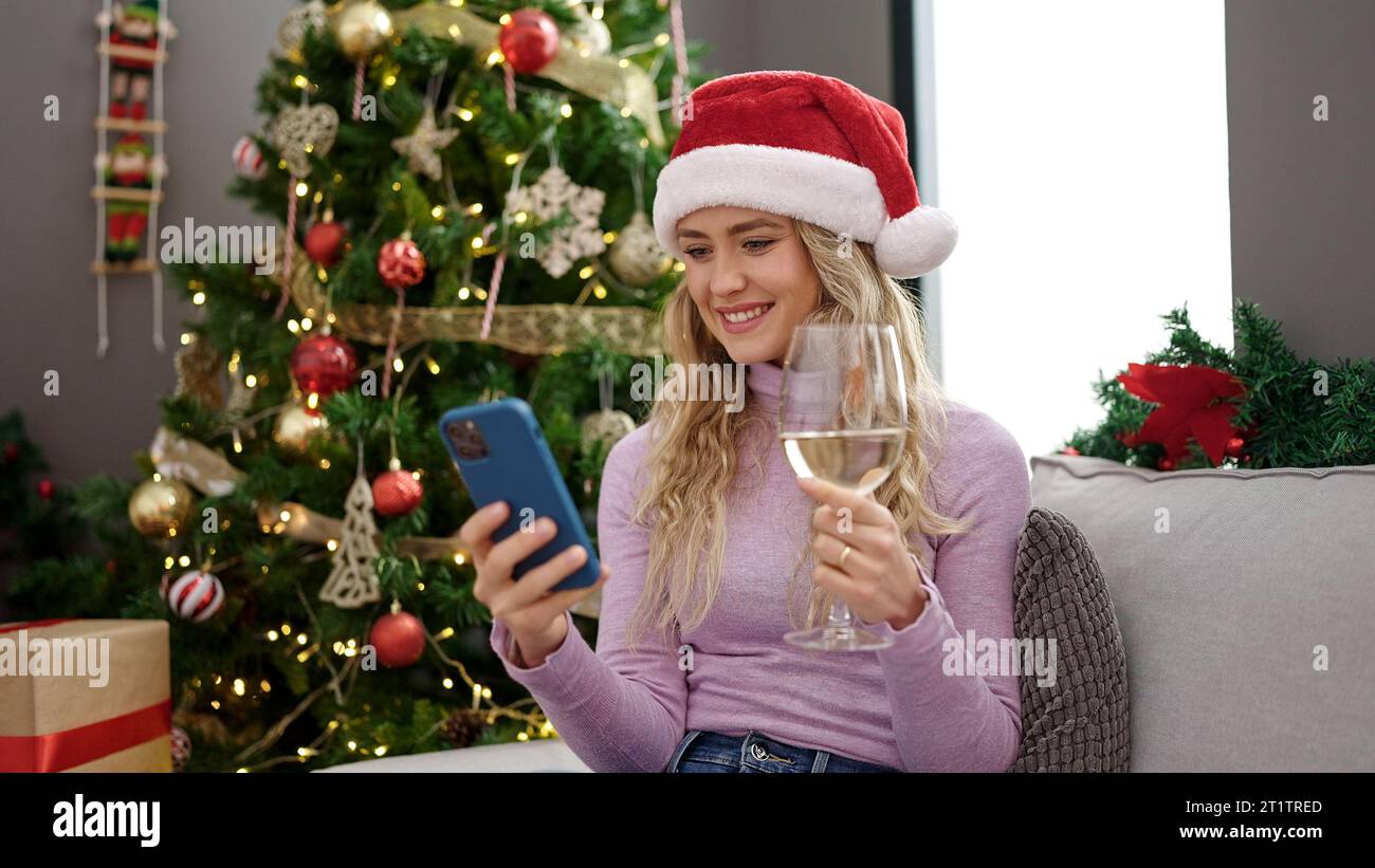 Giovane bionda che festeggia il natale con lo smartphone bevendo vino a casa Foto Stock