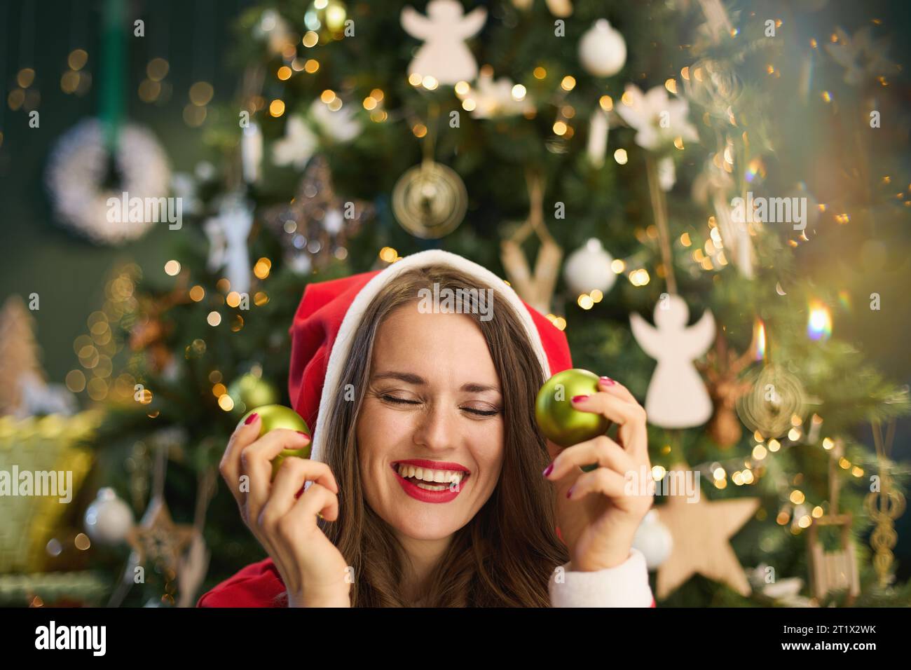 Natale. Casalinga moderna sorridente con cappello di Babbo Natale e palla di Natale vicino all'albero di Natale nella casa moderna. Foto Stock