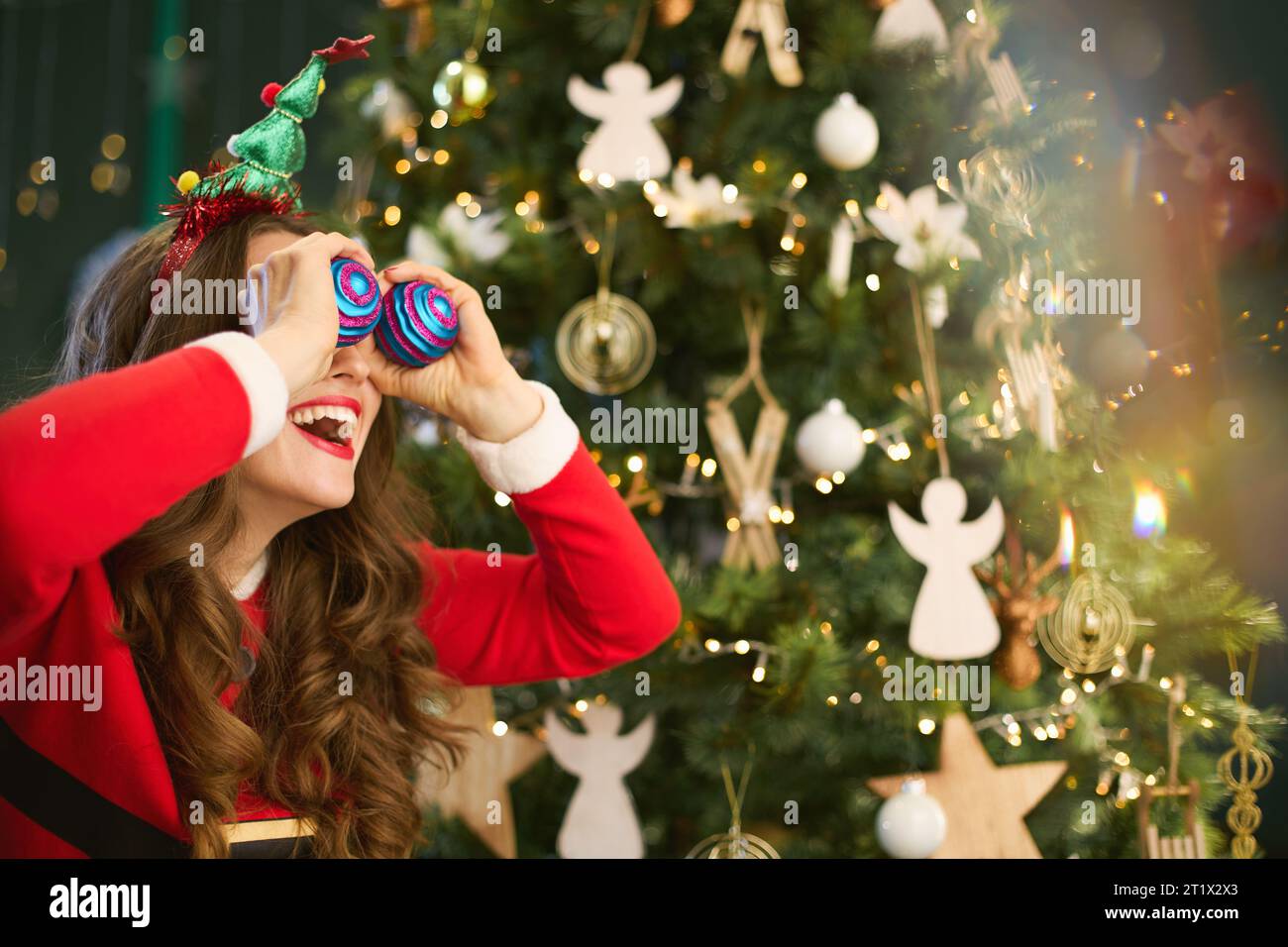 Natale. Elegante e sorridente casalinga di mezza età nel moderno soggiorno che usa le palle di Natale come occhi vicino all'albero di Natale. Foto Stock