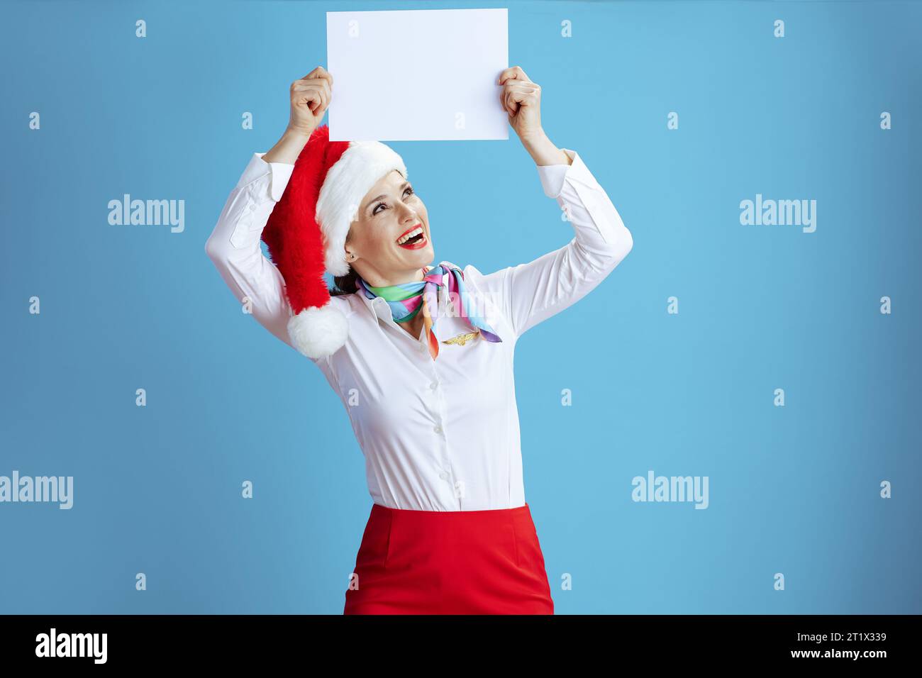 Simpatica ed elegante hostess donna su sfondo blu in uniforme con cappello di Babbo Natale che mostra un foglio di carta a4 vuoto. Foto Stock