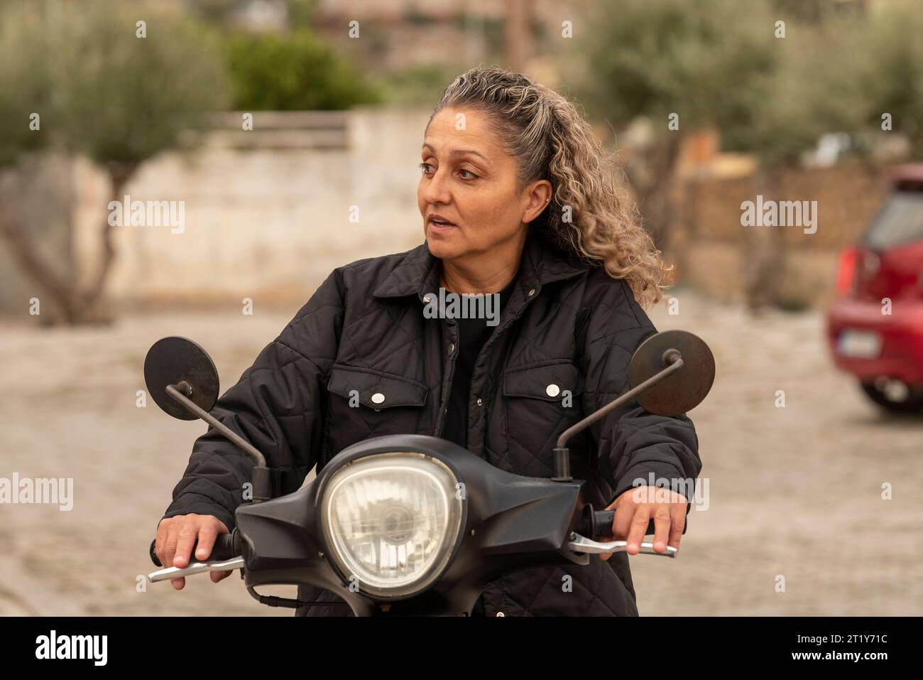 Malia, Creta, Grecia. 03.10.2023. Donna cretese con i capelli ricci e indossa una giacca in camicia a cavallo di uno scooter. Foto Stock