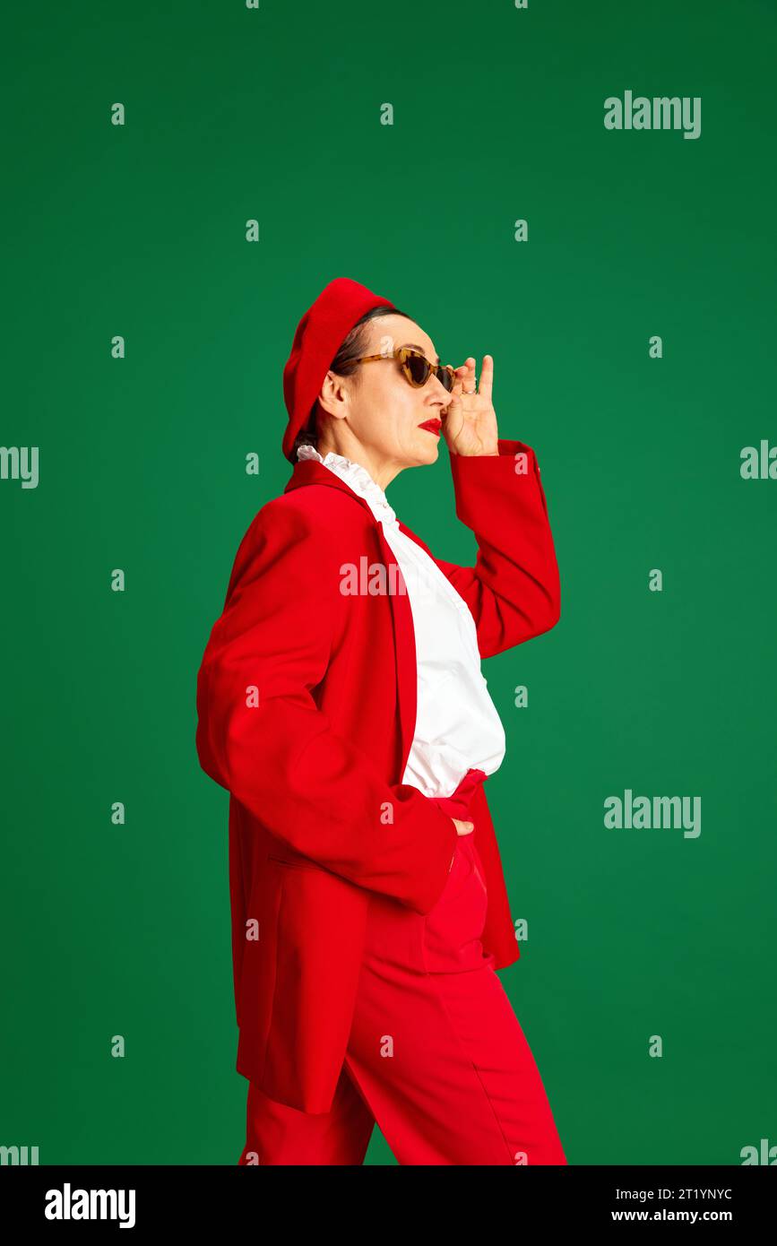 Vista laterale di una donna elegante degli anni '60, che indossa un abito e un berretto rosso brillante ed elegante, in posa sullo sfondo verde dello studio Foto Stock